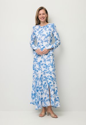 Uśmiechnięta kobieta w białej, długiej sukience maxi z niebieskim kwiatowym wzorem i rozcięciem z przodu, nosząca beżowe sandały na obcasie na otwartym palcu, na jednolitym tle.