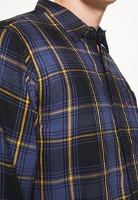 Chemise à carreaux bleu marine, noir et jaune ; dotée d'un col boutonné et d'une poche poitrine. Confectionnée en tissu texturé avec une coupe ajustée.