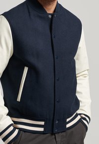 Mörk marinblå jacka i ullblandning med krämfärgade läderärmar, ribbade mudd och krage, tryckknappsstängning och sidofickor. Randig nederdel.