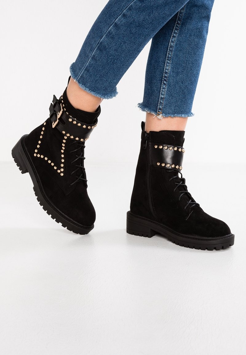 Bottines en daim noir avec clous dorés, brides à boucles, lacets et fermetures éclair latérales portées avec un jean court bleu sur un sol blanc.
