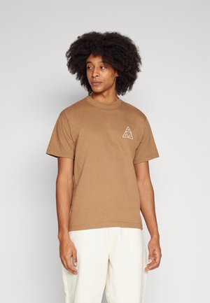 Camiseta de algodón marrón con mangas cortas, que presenta un diseño geométrico en forma de triángulo blanco en el lado izquierdo del pecho. El modelo lleva pantalones de un color claro.