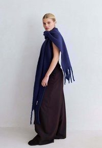 Marineblauwe wollen sjaal met rafelige randen, gedrapeerd over een zwarte top, met een zachte textuur en een lang, vloeiend ontwerp tegen een neutrale achtergrond.