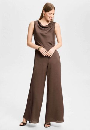 Vrouw draagt een mouwloze bruine jumpsuit met brede pijpen en open hakken, glimlacht met gevouwen handen voor zich tegen een effen achtergrond.