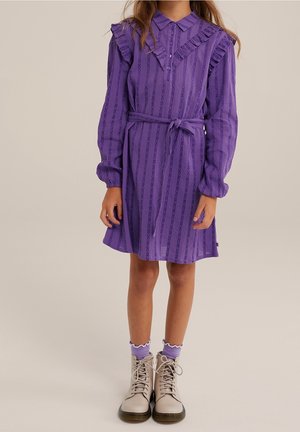 Kind trägt ein langärmliges lila Kleid mit Rüschenbesatz und Gürtel, hellviolette Socken und beige Schnürstiefel, steht vor neutralem Hintergrund.