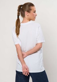 Femme avec une queue de cheval tressée, portant un t-shirt blanc uni à manches courtes et un pantalon foncé, debout, les mains jointes dans le dos.