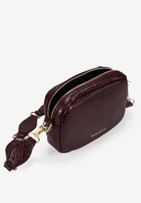 Pequeña bolsa cruzada de cuero texturizado de color burdeos oscuro con cremalleras doradas, correa desmontable y logo "BOSANOVA" en el frente.