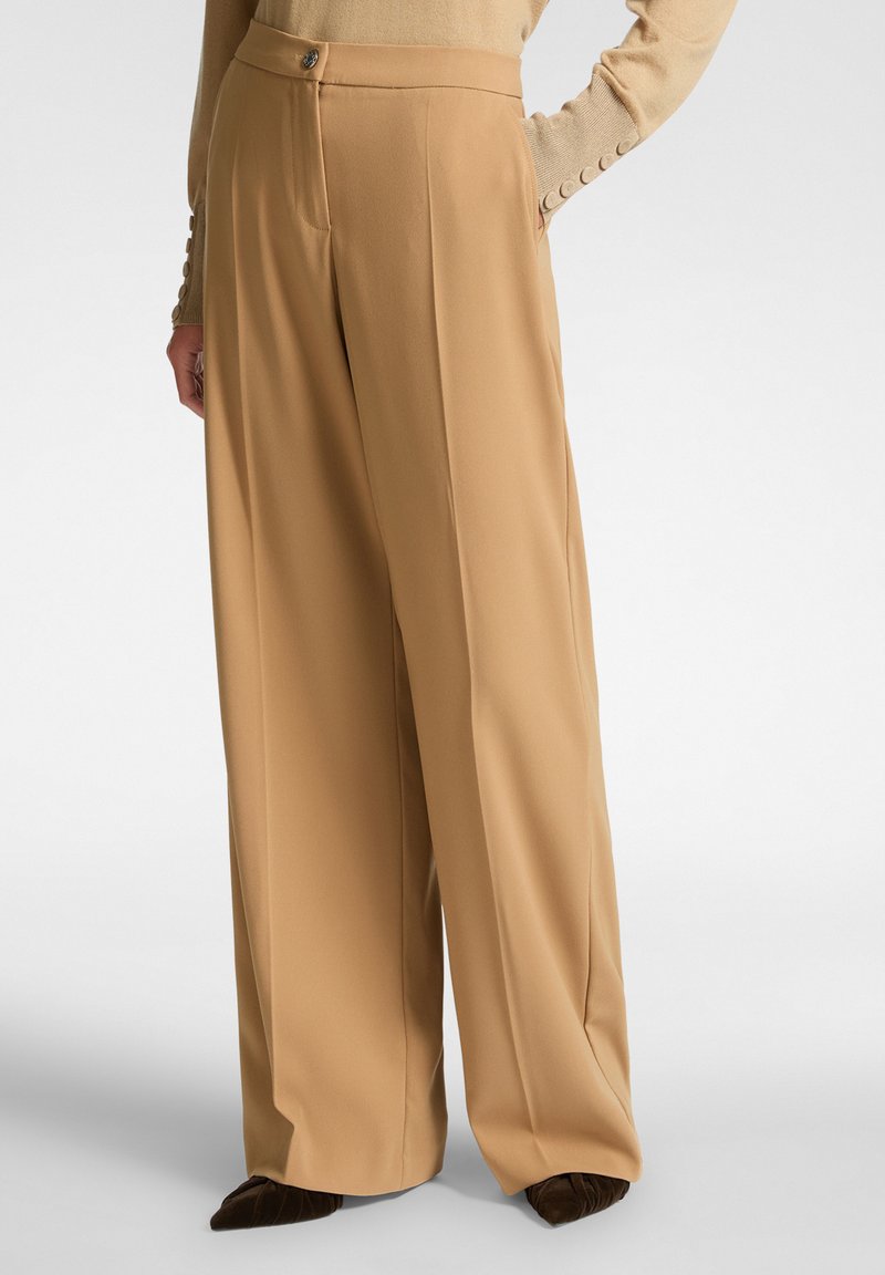 Tan vida byxor med en slät textur, skräddarsydd passform, framfåra och en knappstängning, ihop med en beige topp och mörka skor.