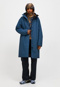 Waterdichte blauwe jas met een capuchon, voorzien van een ritssluiting en zakken, gelaagd over een gebreid patroontrui en zwarte broek.