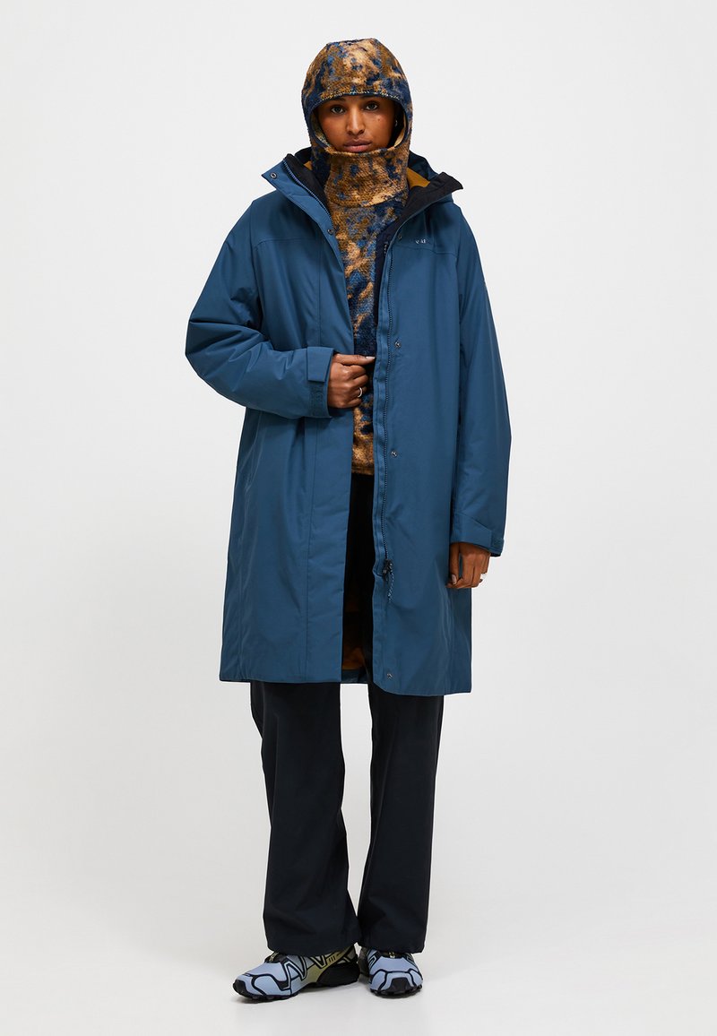 Waterdichte blauwe jas met een capuchon, voorzien van een ritssluiting en zakken, gelaagd over een gebreid patroontrui en zwarte broek.
