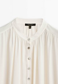 Blouse blanche avec col rond, cinq boutons sur le devant, détails de tissu froncés autour des épaules, et étiquette de marque noire à l’intérieur du col.