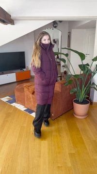 Lang burgunderfarget vattert jakke med høy krage, to frontlommer, knappelukking, brukt over svarte bukser og kraftige svarte sko. Innendørs miljø.