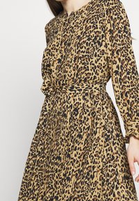 Vestido de estampado de leopardo en tono tostado, con mangas largas, escote redondo y cintura ajustada con un detalle de lazo. La tela plisada crea textura.