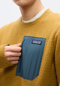 Άτομο που φοράει υφασμένο πουλόβερ μουσταρδί με μπλε φερμουάρ στην τσέπη στο στήθος και ετικέτα λογότυπου Patagonia, τραβώντας το φερμουάρ με το χέρι.