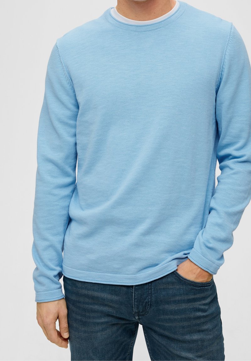 Man wearing light blue long-sleeve crew neck sweater and dark blue jeans, with one hand in pocket and the other relaxed by side.