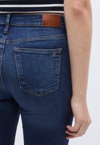 Sötétkék farmernadrág, testhezálló szabással, a derékrészen márkajelzett bőrpáccal és egy kis logóval a hátsó zsebnél.