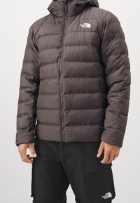 Donkergrijze donsjas met kap, doorgestikt ontwerp, ritssluiting aan de voorkant en logo op de linkerborst. Gestructureerd materiaal met geïsoleerde delen.