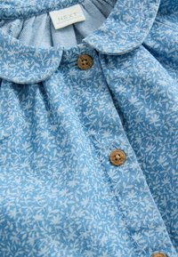 Blouse bleu clair avec imprimé floral, devant plis et col arrondi. Dotée de boutons en bois et d'un tissu texturé.