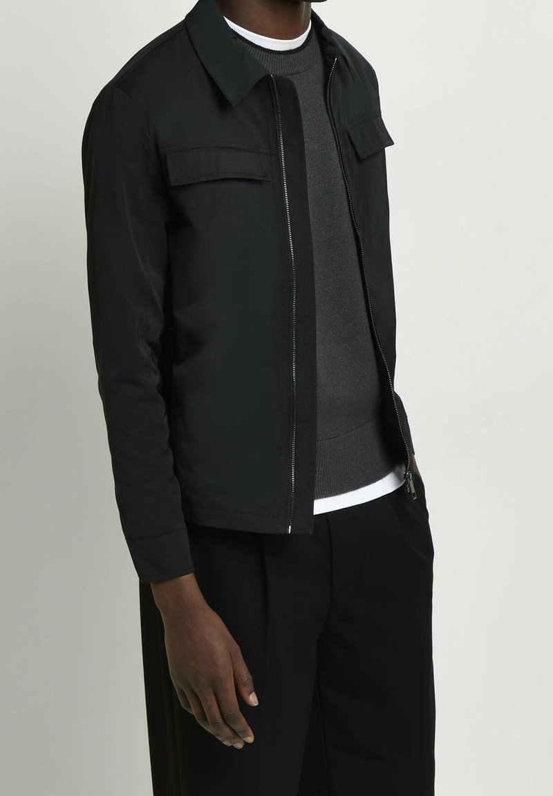 Homme portant une veste zippée noire par-dessus un pull gris et un sous-vêtement blanc, avec un pantalon noir, debout devant un fond clair uni.