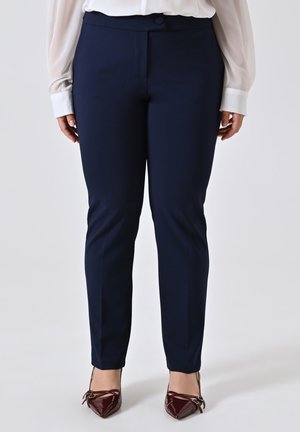 Donna che indossa pantaloni classici attillati blu navy abbinati a una camicia bianca e scarpe con punta in vernice bordeaux e cinturini alla caviglia.