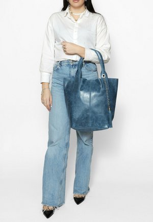 Blue faux leather tote bag with two handles, a smooth finish, and a decorative chain with a tassel. Styled with a white shirt and light jeans.