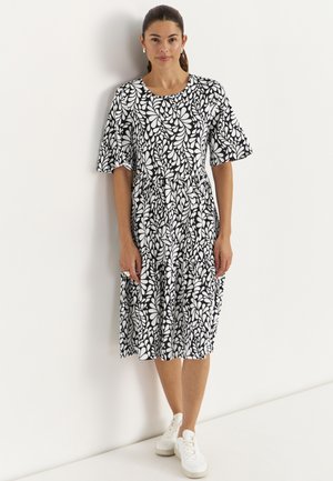 Kvinde med håret bundet tilbage iført sort og hvid midikjole med bladmønster og hvide sneakers, stående foran en hvid væg.