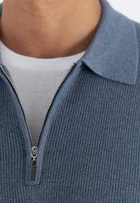 Suéter azul de canalé con frente con cremallera, que presenta un cuello claro y una camiseta interior blanca en contraste. Textura suave con cremallera metálica.