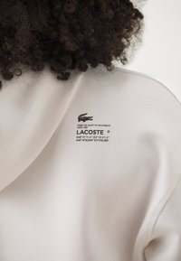 Hanorac bej deschis cu logo-ul Lacoste și text. Fabricat dintr-un material moale, cu o textură netedă. Include coordonate imprimate pe spate.