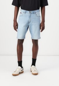 Bermudas de ganga azul claro com corte reto, bordas rematadas, cinco bolsos e fecho de botão, usadas com ténis brancos e pretos.