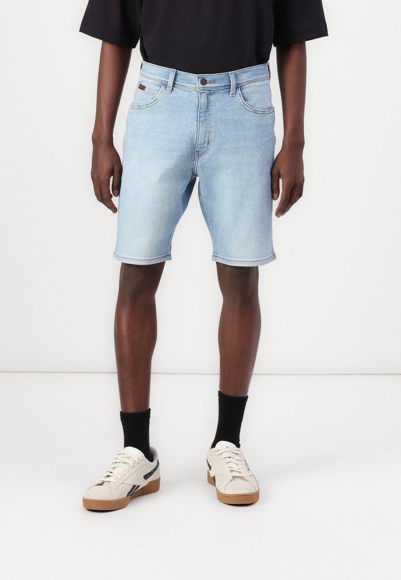 Bermudas de ganga azul claro com corte reto, bordas rematadas, cinco bolsos e fecho de botão, usadas com ténis brancos e pretos.