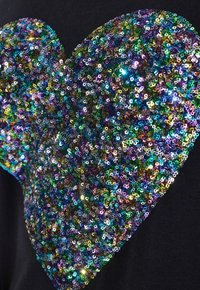 Vêtement noir présentant un grand motif de cœur composé de sequins multicolores dans des teintes de vert, bleu, violet et or, avec une texture brillante.