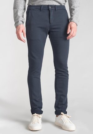 Navy cotton slim-fit pants with front pockets and a button closure, paired with white sneakers, showcasing a casual, modern look.