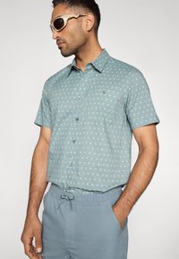 Πουκάμισο με κοντό μανίκι και κουμπιά, σε ανοιχτό μπλε με μικρές μπεζ πουά, με τσέπη στο στήθος και χαλαρή εφαρμογή. Συνδυάζεται με ταιριαστά σορτς.