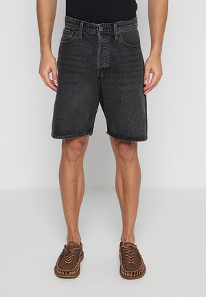 Mand iført sorte denimshorts med fem lommer og brune vævede snøresko, stående mod en ensfarvet lys grå baggrund.