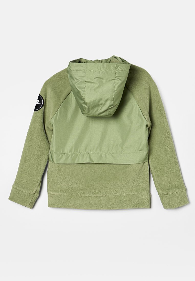 Veste en polaire verte avec capuche, présentant une partie supérieure lisse, une partie inférieure texturée et un logo noir rond sur la manche gauche.
