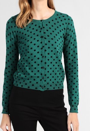 Kvinde iført grøn cardigan med lange ærmer med sorte polkaprikkker og sorte knapper, kombineret med sorte bukser.