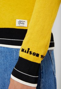 Żółty sweter z dzianiny z czarno-białymi, prążkowanymi mankietami. Na piersi znajduje się metka z napisem "Maison Scotch" oraz logo na rękawie.