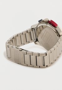 Ασημένιο μεταλλικό μπρασελέ ρολογιού με πόρπη, συνδεδεμένο στο σώμα του ρολογιού με στρογγυλή πλάτη και κουμπιά κόκκινου και μαύρου χρώματος.