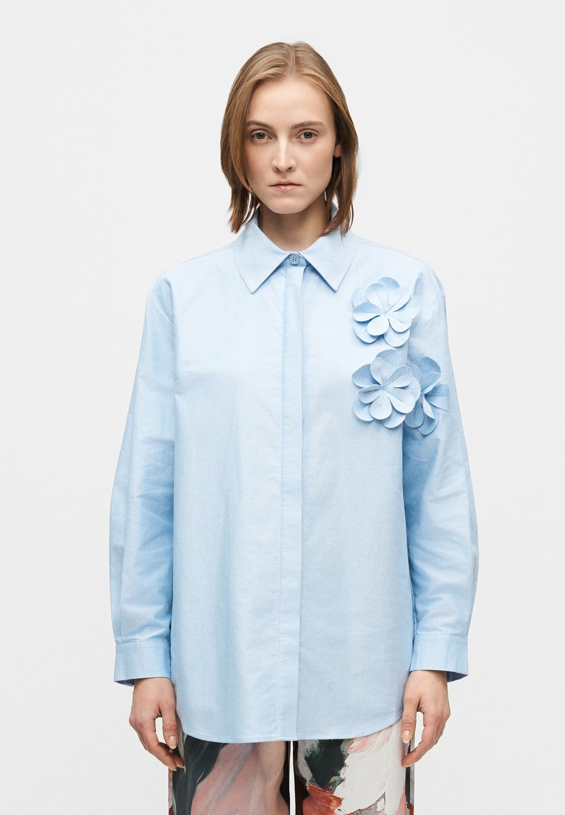 Cămașă bleu deschis din bumbac, cu aplicatii florale tridimensionale pe partea stângă și mâneci lungi.