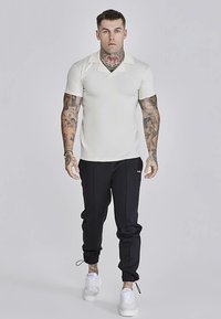 Vit pikétröja med platt krage, kombinerad med svarta avsmalnande joggers och vita sneakers. Tatueringar syns på armarna.