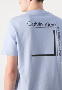 Lyseblå bomulds T-shirt med sort tekst og grafisk design på ryggen, som står "Calvin Klein Established Since 1978."