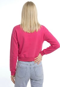 Blond persoon draagt een roze shirt met lange mouwen en lichtblauwe spijkerbroek, staat met de rug naar de camera en rechterhand op de heup.
