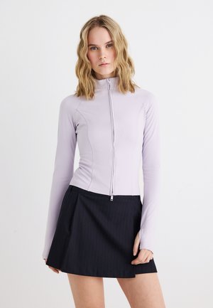 Naine laineliste blondide juustega, kandes helelillat pika varrukaga tõmblukuga pusa ja musta voltidega seelikut valgel taustal.