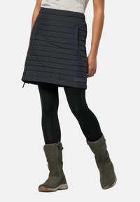 Donkergrijze geïsoleerde skort met horizontale stiksels, zijzak en een logo. Draagt bij de zwarte leggings en grijze laarzen tot aan de kuiten.