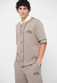 Jauns vīrietis ar cirtainiem matiem, tērpies bežā krāsā Armani Exchange pogājamo krekla un saskaņotu bikses, ar rokām kabatās, skatās uz priekšu.