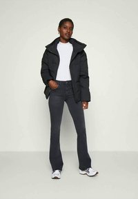 Μαύρο φουσκωτό μπουφάν με ψηλό γιακά, λευκό μπλουζάκι, σκούρο τζιν και λευκά αθλητικά παπούτσια με καφέ α accents. Στάση όρθια μπροστά σε ουδέτερο φόντο.