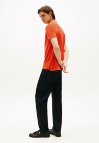 Pomarańczowa koszulka polo, ciemne dżinsy oraz czarne mokasyny. Koszulka ma krótkie rękawy i boczne rozcięcia; dżinsy mają tylne kieszenie i widoczne szwy.