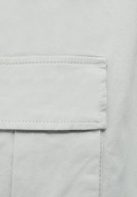 Gros plan sur une poche en tissu gris pâle avec un rabat et des coutures visibles sur un vêtement.