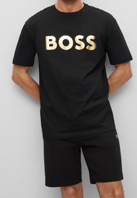 Man i svart T-shirt med stort guld "BOSS"-tryck och svarta shorts mot en grå bakgrund, med händerna bakom ryggen.