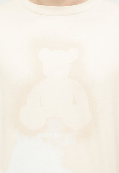 Γραφικό ξεθωριασμένης αρκουδίτσας σε ανοιχτό μπεζ χρώμα πάνω σε ύφασμα εκρού, κεντραρισμένο με στρογγυλή λαιμόκοψη από πάνω.