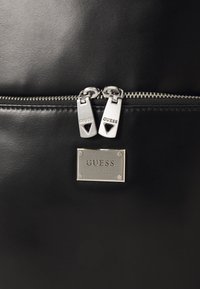 Zaino in pelle nera con zip argentata a doppio cursore, una piastrina metallica con il marchio e una texture liscia.